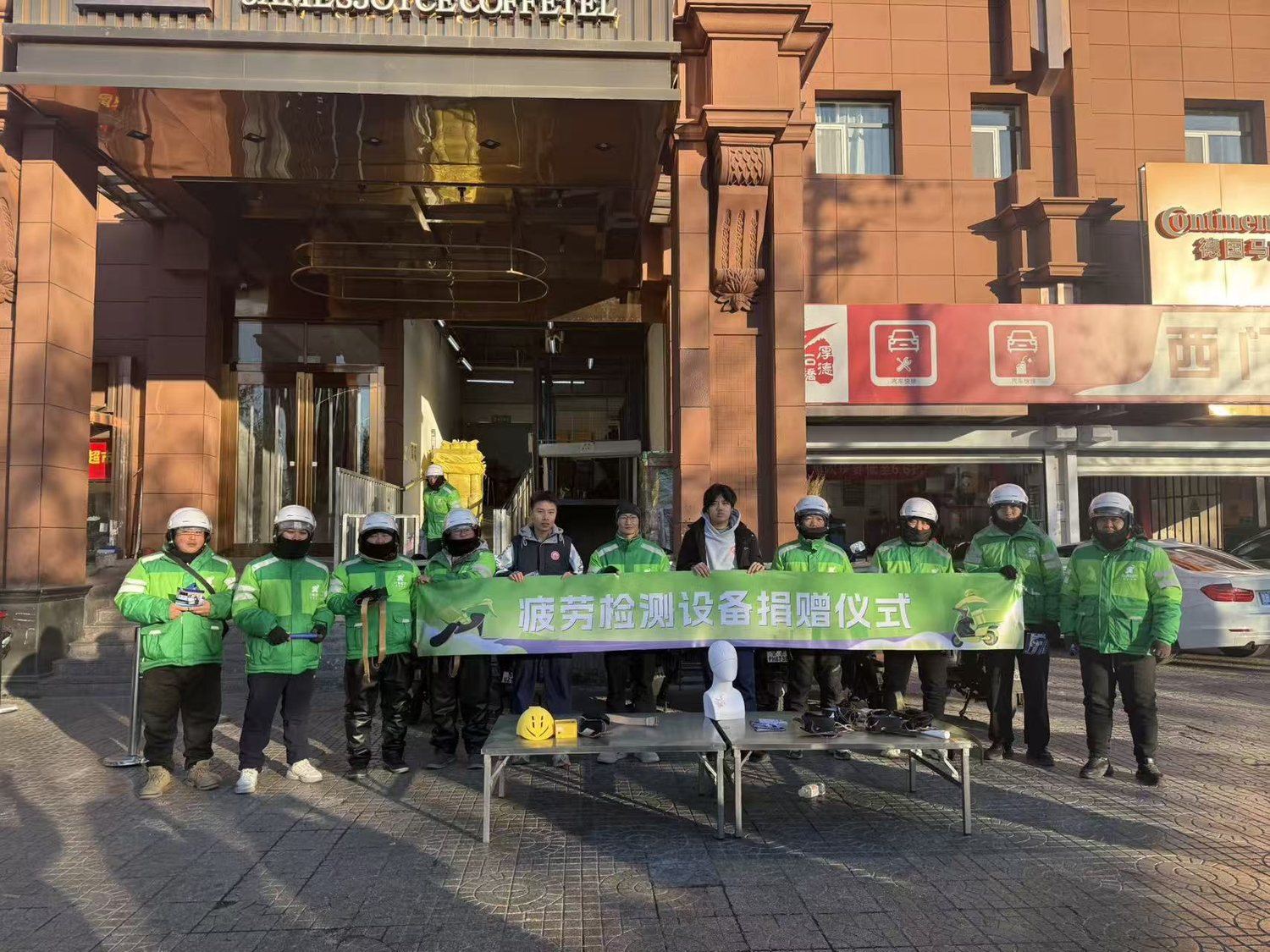 Delivery riders in green uniforms holding a fatigue detection equipment donation banner outside a building.