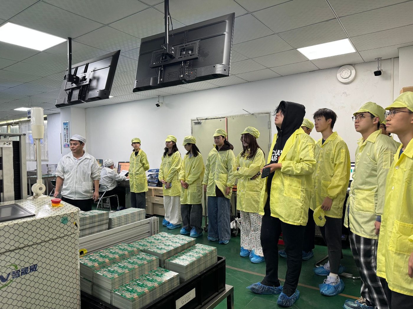 Visitors in cleanroom coats and shoe covers observing an electronics production line with stacked cartons.